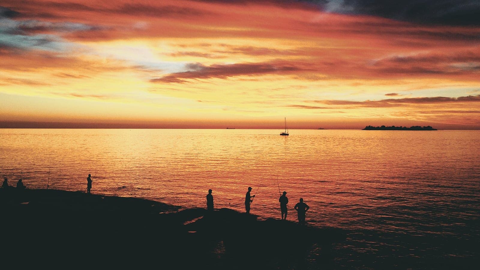 Atardecer anaranjado sobre el Río de la Plata con siluetas de pescadores en la orilla y un velero a lo lejos