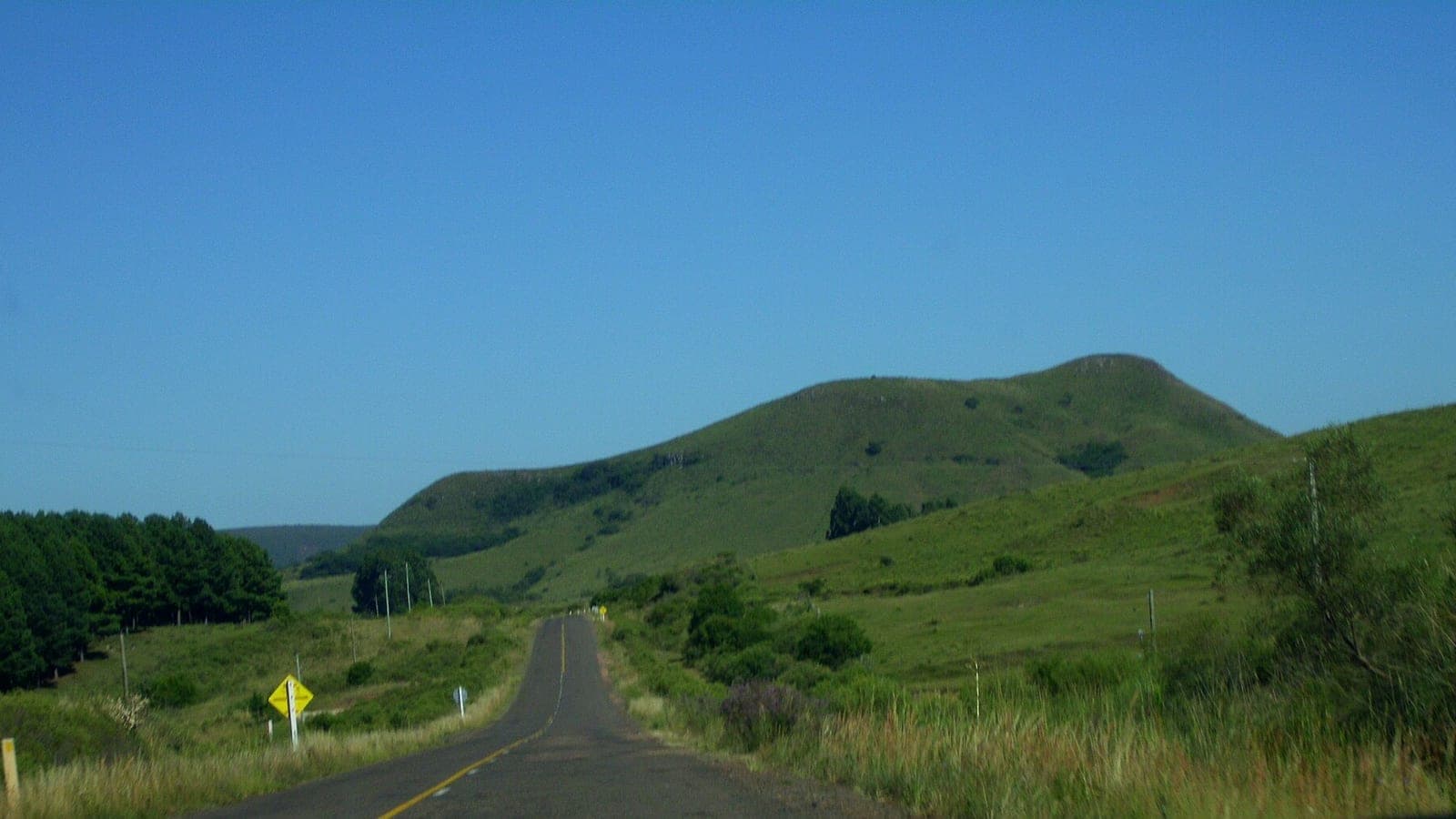 Ruta de asfalto con línea amarilla central que atraviesa cuchillas verdes bajo un cielo despejado