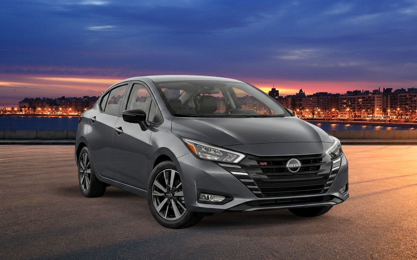 Nissan Versa gris de frente y tres cuartos, con la costa de Montevideo iluminada al anochecer detrás